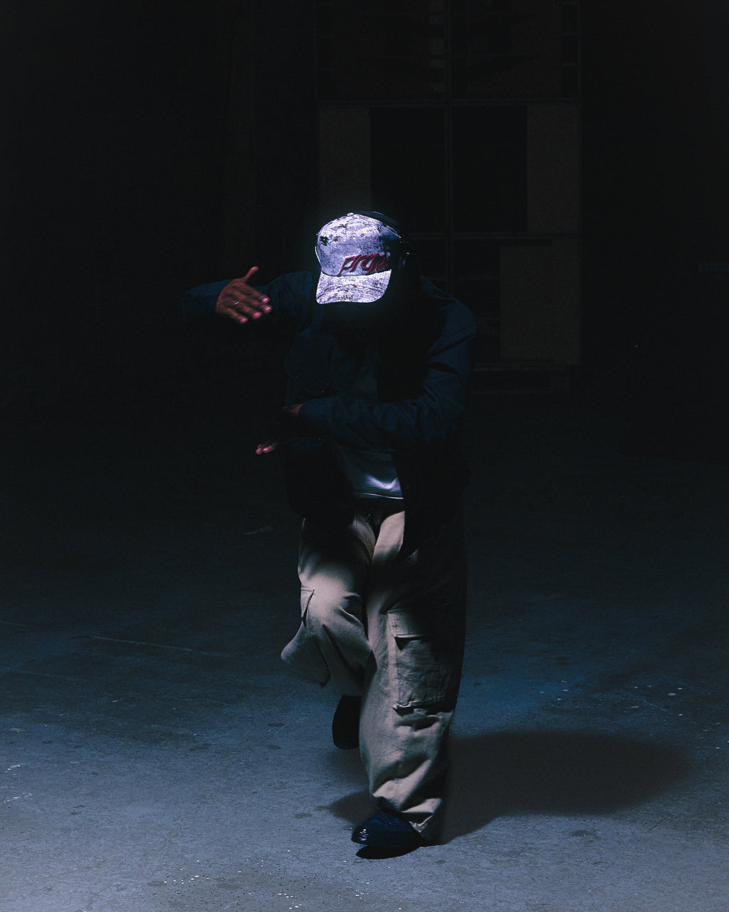 A dancer wearing the ANTIFRAGILE camo strapback hat with a dark outfit and beige pants. They are dancing in an abandoned warehouse.