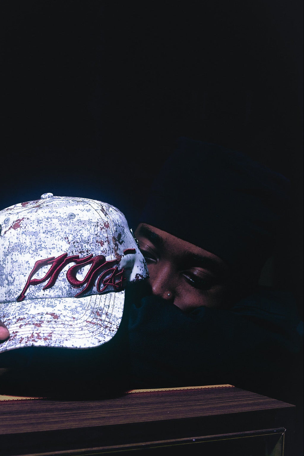Person holding ANTIFRAGILE  maroon red, grey and white camo print strapback hat. He leans on top of a rustic speaker, showcasing the maroon red 3D puff print embroidery.